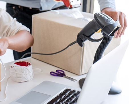 Duas pessoas embalam caixas com laptop, scanner, tesoura e outros itens em cima da mesa.